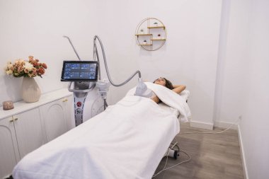 Genç bir kadın tedavi yatağında istilacı olmayan bir vücutla çevrili olarak uzanıyor. Parlak, profesyonel bir sağlık kliniğinde zayıflama ve gençleştirme için gelişmiş ekipmanlarla.