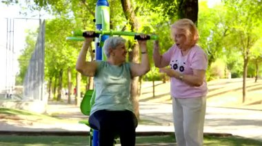 Aktif bir yaşam tarzının keyfini çıkaran iki neşeli son sınıf öğrencisi güneşli bir parkta spor aletleri üzerinde egzersiz yapıyor. Arkadaşı motivasyon ve destek sağlarken bir kadın omuz pres makinesi kullanıyor.