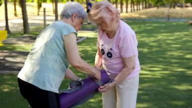 İki yaşlı kadın, güneşli bir parkta mor bir yoga minderini birlikte sarıp emekliliklerinden sonra arkadaşlıklarıyla aktif ve sağlıklı bir yaşam tarzı sergiliyorlar.