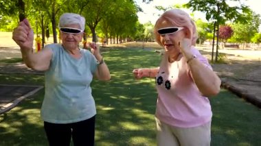 İki neşeli son sınıf öğrencisi güneş gözlüğü takıyor. Parkta mutlu bir şekilde dans ediyorlar. Güneşli bir günde çevrende yeşil ağaçlarla aktif ve sağlıklı emeklilik hayatlarının tadını çıkarıyorlar.