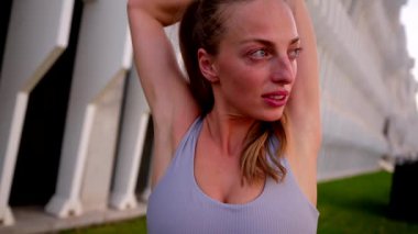 Eğitiminden önce atkuyruğu olan güzel atletik bir kadın günbatımında kollarını, sırtını ve bacaklarını modern bir şehir parkında açıyor.