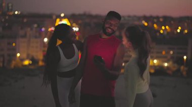 Spor giyim sektöründe çok ırklı bir grup arkadaş çatıda güzel bir şehir manzaralı akıllı bir telefon kullanırken birlikte gülüp sohbet ediyorlar.