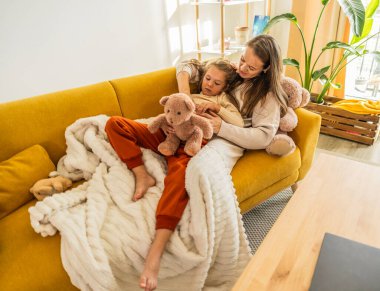 Mother embracing her daughter and teddy bears while relaxing on a comfortable couch at home, enjoying a cozy moment of family bonding and affection in the living room