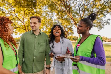 Yansıtıcı yelek giyen çeşitli genç gönüllüler, güneşli bir parktaki bir toplum araştırması sırasında katılımcılarla mutlu bir şekilde etkileşime geçiyor, gülüyor ve bir panoya notlar yazıyorlar.