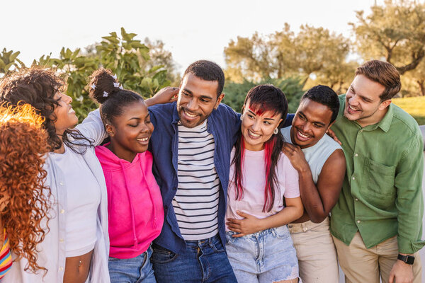 Diverse group of smiling young adults standing close, embracing each other and laughing together, enjoying friendship and togetherness outdoors in natural light
