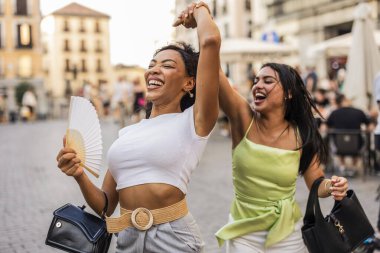 Two cheerful multi ethnic women friends enjoying a fun summer day, laughing and dancing together on a city street, expressing joy, freedom, and friendship