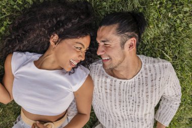 Happy diverse couple lying together on green grass, looking at each other, and laughing in a top down view, representing joy, love, togetherness, and connection outdoors