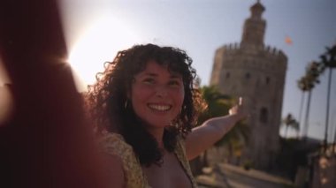 Happy young woman vlogging for social media, pointing at the torre del oro in seville, spain, smiling and blowing a kiss to the camera during a beautiful sunny day with a warm lens flare