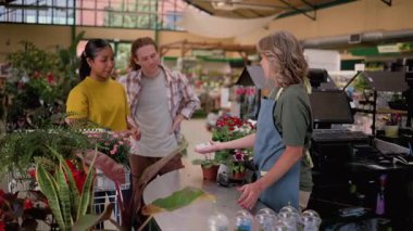 Happy young customer at a flower shop checkout making a contactless payment with a credit card for her purchases, assisted by a smiling female cashier in a bright and lively plant nursery