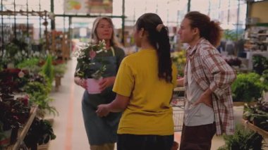 Knowledgeable female employee in an apron helping a diverse young couple select a beautiful potted plant with white flowers while shopping inside a large, bright plant nursery greenhouse