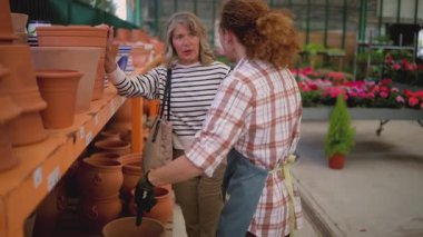 Mature female florist wearing an apron managing plant inventory with a digital tablet at the counter of a flower shop, checking potted petunias and organizing the display for sale