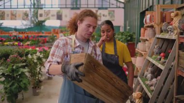 Mature female florist wearing an apron managing plant inventory with a digital tablet at the counter of a flower shop, checking potted petunias and organizing the display for sale