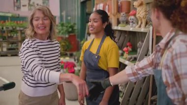 Mature female florist wearing an apron managing plant inventory with a digital tablet at the counter of a flower shop, checking potted petunias and organizing the display for sale