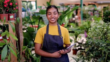 Mature female florist wearing an apron managing plant inventory with a digital tablet at the counter of a flower shop, checking potted petunias and organizing the display for sale