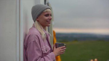 Pembe ceketli ve bereli gülümseyen sarışın kadın elinde dumanı tüten bir kupayla karavanına yaslanıyor ve huzurlu bir kır gezisinin tadını çıkarıyor.