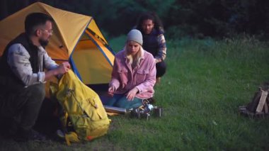 Bir grup farklı arkadaş, gece boyunca huzurlu bir ormanlık alanda kamp kuruyor. Sırt çantasını sarı çadırın yanında açan bir adamla birlikte iki kadın da gece için hazırlanıyor.