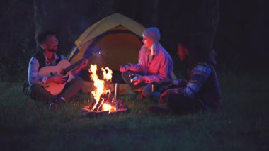 Neşeli çoklu etnik arkadaşlar birlikte yaz gecesinin tadını çıkarıyorlar, kamp ateşi etrafında çimlerin üzerinde oturup, bir adam çadırın önünde akustik gitar çalarken şarkılar söylüyorlar.