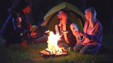 Çok ırklı arkadaşlar gece kamp gezisinin tadını çıkarıyorlar. Şenlik ateşinin etrafında çimlerin üzerinde oturup gülüyorlar ve çubuklarda marşmelov kızartıyorlar. Sıcak ışıkta görünen bir çadırla.