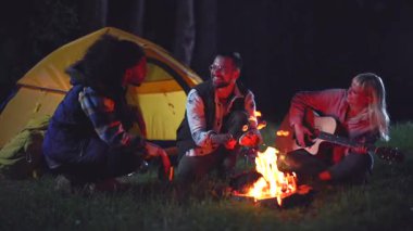 Neşeli çeşitli arkadaşlar gece ormanda şenlik ateşinin etrafında dinlenirken, bir kadın akustik gitar çalıyor ve diğerleri birlikte şarkı söylüyor, açık hava maceralarının tadını çıkarıyorlar.