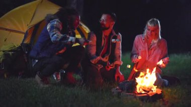 Üç çok kültürlü arkadaş çimlerin üzerinde birlikte oturur, arka planda sarı bir çadırla gece kamp yaparken parlayan bir kamp ateşinde gülerek şekerleme kızartırlar.