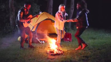 Bir grup çok ırklı genç arkadaş gece ormanda kamp yaparken, bir adam sarı bir çadırın yanında akustik gitar çalarken, şenlik ateşi etrafında şarkı söyleyip dans ederken eğleniyorlar.