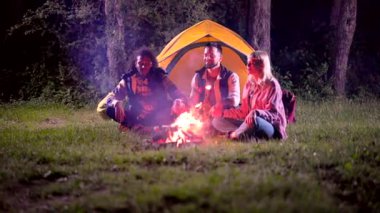 Üç farklı arkadaş, iki erkek ve bir kadın, gece ormanda kamp yapıyorlar, gülüyorlar ve konuşuyorlar, sıcak bir şenlik ateşinin etrafında oturuyorlar, parlak sarı çadırlarının yanında.
