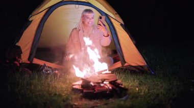 Neşeli bayan gezgin çadırın içinde oturuyor, akıllı telefonunu video görüşmesi için kullanıyor ve termo bardaktan içiyor, macerasını gece çıtırdayan bir kamp ateşinin ışığında paylaşıyor.
