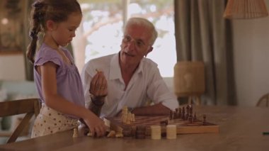 Bilge kıdemli bir adam, mutlu torununa satranç oynamayı öğretiyor, kuralları anlatıyor ve sıcak, güneşli bir oturma odasında, ahşap bir masada otururken ona parçaları gösteriyor.