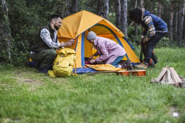 Çeşitli arkadaşlar kamp ateşinin yanında turuncu çadırlarını ve kamp malzemelerini hazırlıyorlar ve yemyeşil bir ormanda akustik gitar çalıyorlar. Açık hava macerasının ve doğa macerasının tadını çıkarıyorlar.
