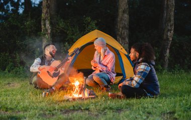 Ormanda kamp yapan üç farklı arkadaş canlı bir kamp ateşinin etrafında oturuyor, biri gitar çalıyor ve neşeyle şarkı söylüyor diğerleri ise açık havada dinleyip bir an için bağlantı kuruyorlar.