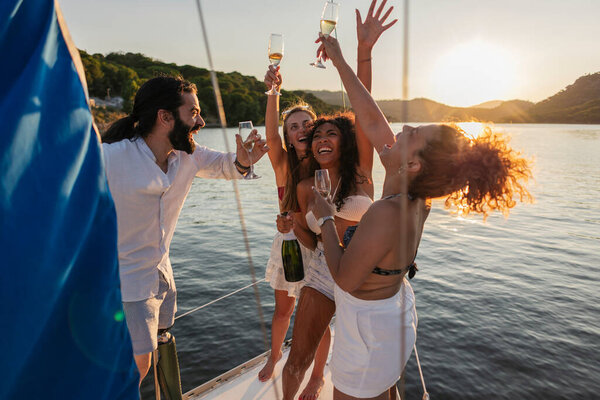 Group of excited friends enjoying a celebration on a yacht, raising glasses of champagne for a toast while watching the beautiful golden sunset over the water