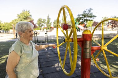 Beyaz saçlı yaşlı bir kadın açık hava fitness parkında kol döndürme makinesi kullanırken gülümsüyor, sağlıkla ve sağlıklı bir yaşam tarzıyla aktif olarak meşgul oluyor.