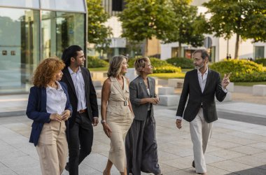 Modern bir şehir meydanında birlikte yürüyen, aktif olarak fikirleri tartışan ve projeleri üzerinde işbirliği yapan, takım çalışmalarını ve profesyonel ağları destekleyen beş iş adamından oluşan çeşitli bir grup.