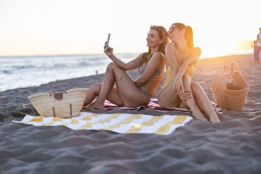 Okyanus kıyısında bir yaz akşamının keyfini çıkaran iki gülen arkadaş, bir kadın cep telefonuyla selfie çekerken, kumsalda çizgili bir havlunun üzerinde oturuyorlar.