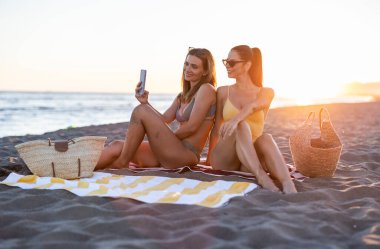 Gün batımında kumlu bir sahilde oturan iki neşeli genç kadın, akıllı telefonlarıyla selfie çekiyorlar, yaz tatillerinde mutlu anlarını paylaşıyorlar.