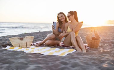 Gün batımında kumlu bir plaj havlusunda oturan iki gülümseyen kadın arkadaş, akıllı bir telefonla özel bir anı kaydediyor, deniz kenarında rahatlatıcı bir yaz tatilinin tadını çıkarıyorlar.
