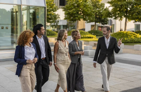 Modern bir şehir meydanında birlikte yürüyen, aktif olarak fikirleri tartışan ve projeleri üzerinde işbirliği yapan, takım çalışmalarını ve profesyonel ağları destekleyen beş iş adamından oluşan çeşitli bir grup.
