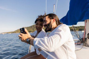 Neşeli çift, güneşli bir yelkenlide güneşli bir tatil gününün tadını çıkarıyor, birlikte anıları yakalıyor ve cep telefonlarıyla okyanusu keşfederken eğleniyorlar.