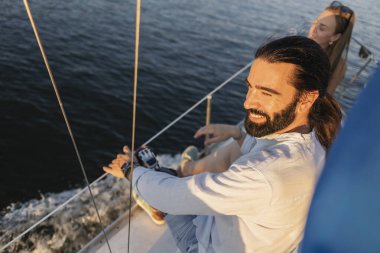 Sakallı ve uzun saçlı bir adam yelkenlide dinlenirken gülümsüyor, arka planda bir kadınla sakin bir okyanusta altın saatlik yolculuğun tadını çıkarıyor, eğlence ve özgürlüğü simgeliyor.