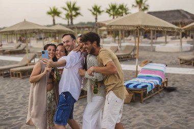 Mutlu çoklu etnik gruplardan oluşan bir grup gülüyor ve kumlu bir sahilde gün batımında selfie çekiyor kaygısız bir yaz tatili partisinde, birlikte neşeli anılarını paylaşıyorlar.