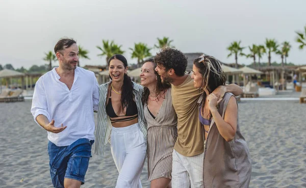 Neşelerini ve beraberliklerini ifade eden bir grup arkadaş gün batımında kumlu bir sahilde kol kola yürürken gülüyorlar yaz tatillerinin ve kaygısız bir yaşam tarzının tadını çıkarıyorlar.