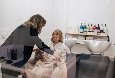 Kuaför, neşeli, olgun bir kadını modern bir kuaförde saç yıkama deneyimi için hazırlıyor, rahatlatıcı ve profesyonel bir atmosfer yaratıyor.