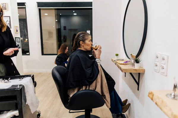 Saçlarını yıkatan, koruyucu pelerinli bir sandalyede oturan, modern bir kuaförde kahve içerken bir anlık rahatlama ve özgüvenin tadını çıkaran bir kadın.