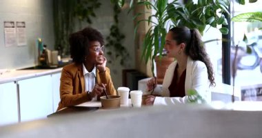 Modern bir ofis dinlenme odasında masada oturan iki farklı iş kadını konteynırlardan yemek yiyor ve öğle yemeği arasında arkadaşça, gülümseyerek sohbet ediyorlar.