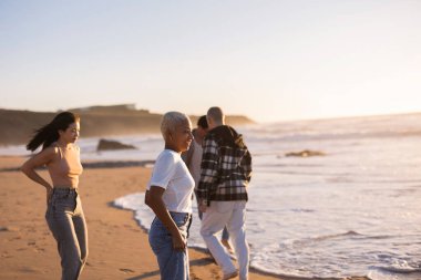 Gün batımında kumlu bir plajda yürüyüş yapan çeşitli arkadaş grupları. Okyanus dalgaları yavaşça kıyıya vuruyor. Arkadaşlık, birliktelik ve açık hava eğlencesi kavramlarını vurguluyor.