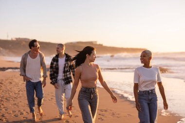 Günbatımında kumlu bir sahil boyunca çıplak ayakla yürüyüp, arkadaşlığı, özgürlüğü ve okyanus dalgaları yakınındaki mutluluğu kucaklayan çeşitli gülen arkadaşlar grubu.