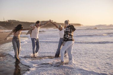 Dört farklı arkadaş el ele tutuşuyor, gülüyor ve kumlu bir kumsalda köpüklü okyanus dalgalarında oynuyorlar. Güzel bir altın saat gün batımında, dostluk ve mutluluğu resmediyorlar.