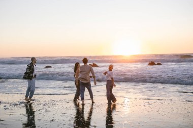 Genç arkadaşlar kumlu bir plajda canlı bir altın saat günbatımının tadını çıkarıyorlar, çıplak ayakla gülüyorlar ve kaygısız bir yaz kaçamağı sırasında ılık okyanus dalgalarında yürüyüp oynuyorlar.