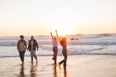 Bir grup farklı arkadaş, güzel turuncu bir günbatımında sahilde özgürlüğü ve mutluluğu kutluyor, suda yalınayak yürüyor ve okyanus dalgalarının yanında oynuyorlar.