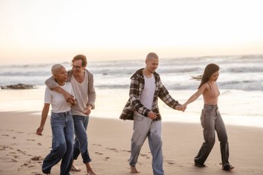 Gün batımında kumlu bir sahilde el ele tutuşan ve çıplak ayakla yürüyen çok nesildir aile, okyanus kenarında sıcak ve kaygısız bir tatilin tadını çıkarıyor ve gülüyor.
